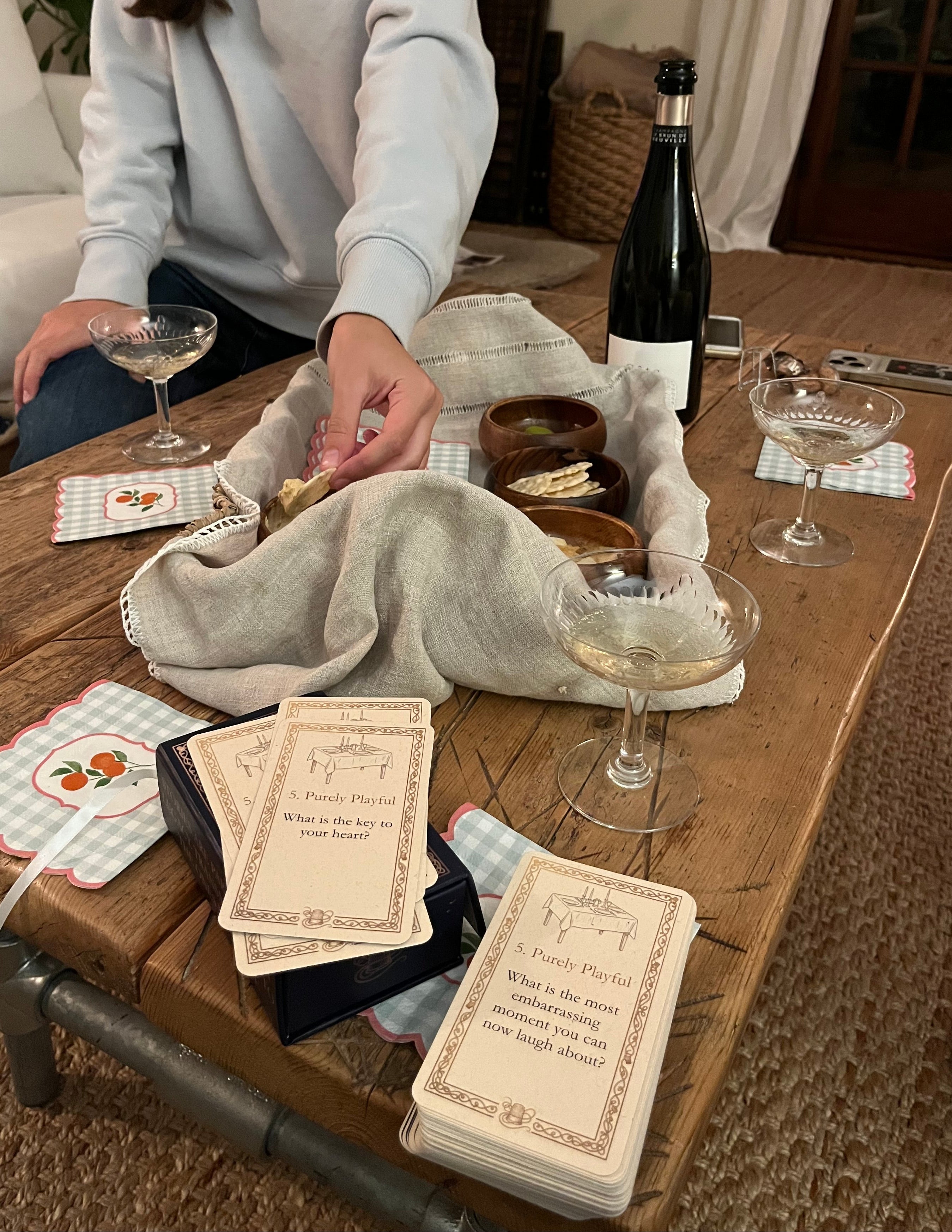 A person in a light blue sweatshirt is seated at a rustic wooden table in a cozy indoor setting, arranging a game setup with cards, small bowls, a champagne bottle, and champagnes glasses.