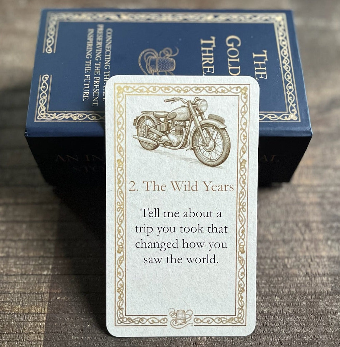 A card with question 'Tell me about a trip you took that changed how you saw the world.' in category '2. The Wild Years' from 'The Golden Thread' deck is displayed in front of its dark blue box on a dark wooden tray.