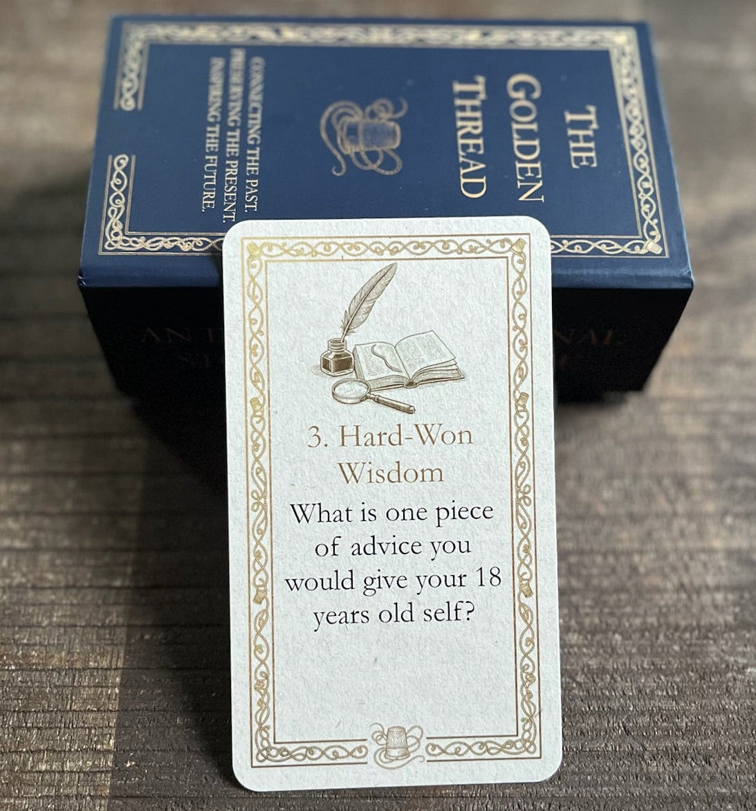 A card with question 'What is one piece of advice you would give your 18 years old self?' in category '3. Hard-Won Wisdom' from 'The Golden Thread' deck is displayed in front of its dark blue box on a dark wooden tray.