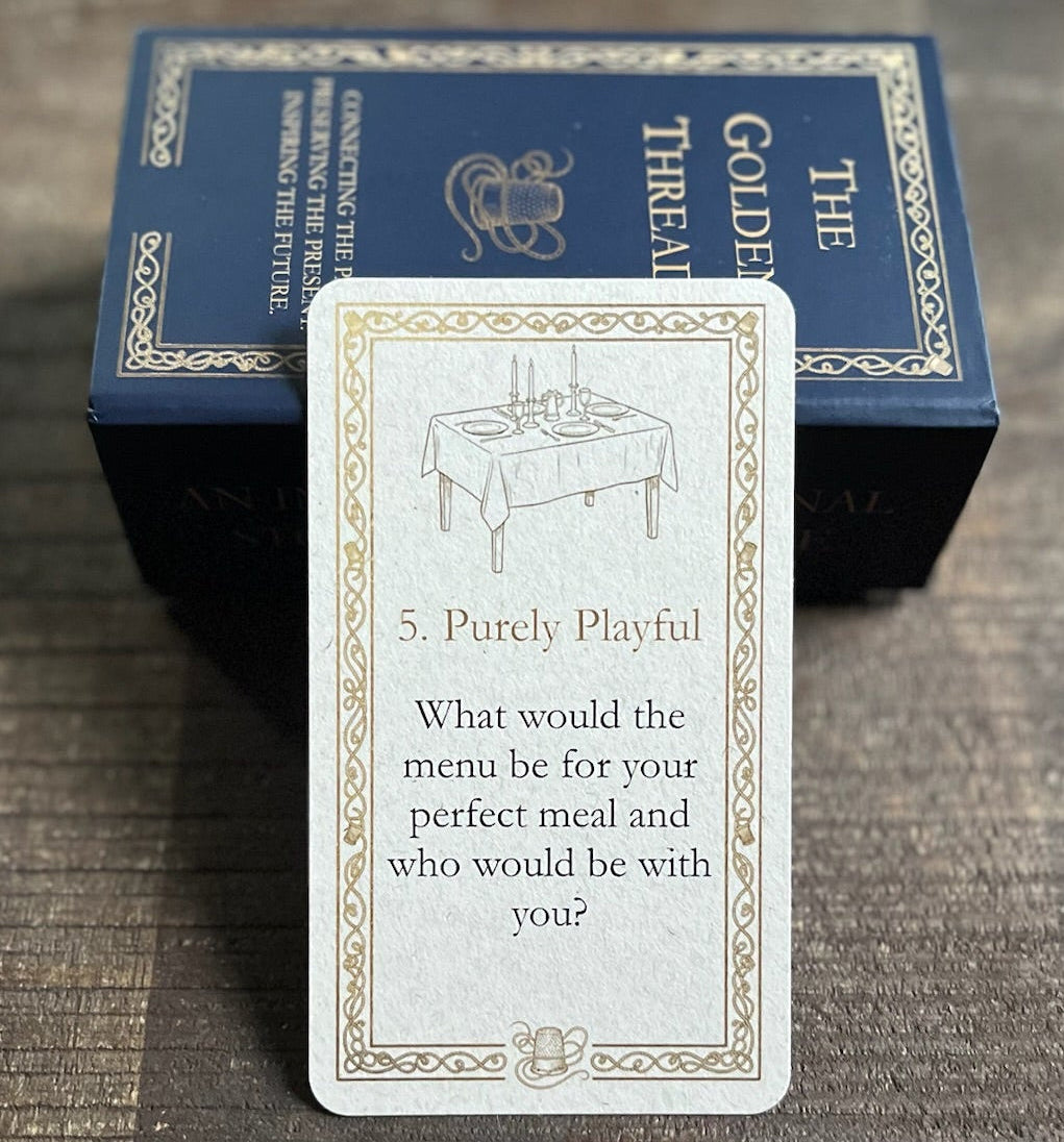 A card with question 'What would be the menu for your perfect meal and who would be with you?' in category '5. Purely Playful' from 'The Golden Thread' deck is displayed in front of its dark blue box on a dark wooden tray.